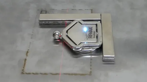Process video showing a fiber laser machine engraving a 360-degree rotating house keychain. Detailed view of the laser marking RE/MAX Québec logos, agent contact information, and a functional QR code on a metal surface.